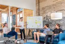 three men sitting while using laptops and watching man beside whiteboard